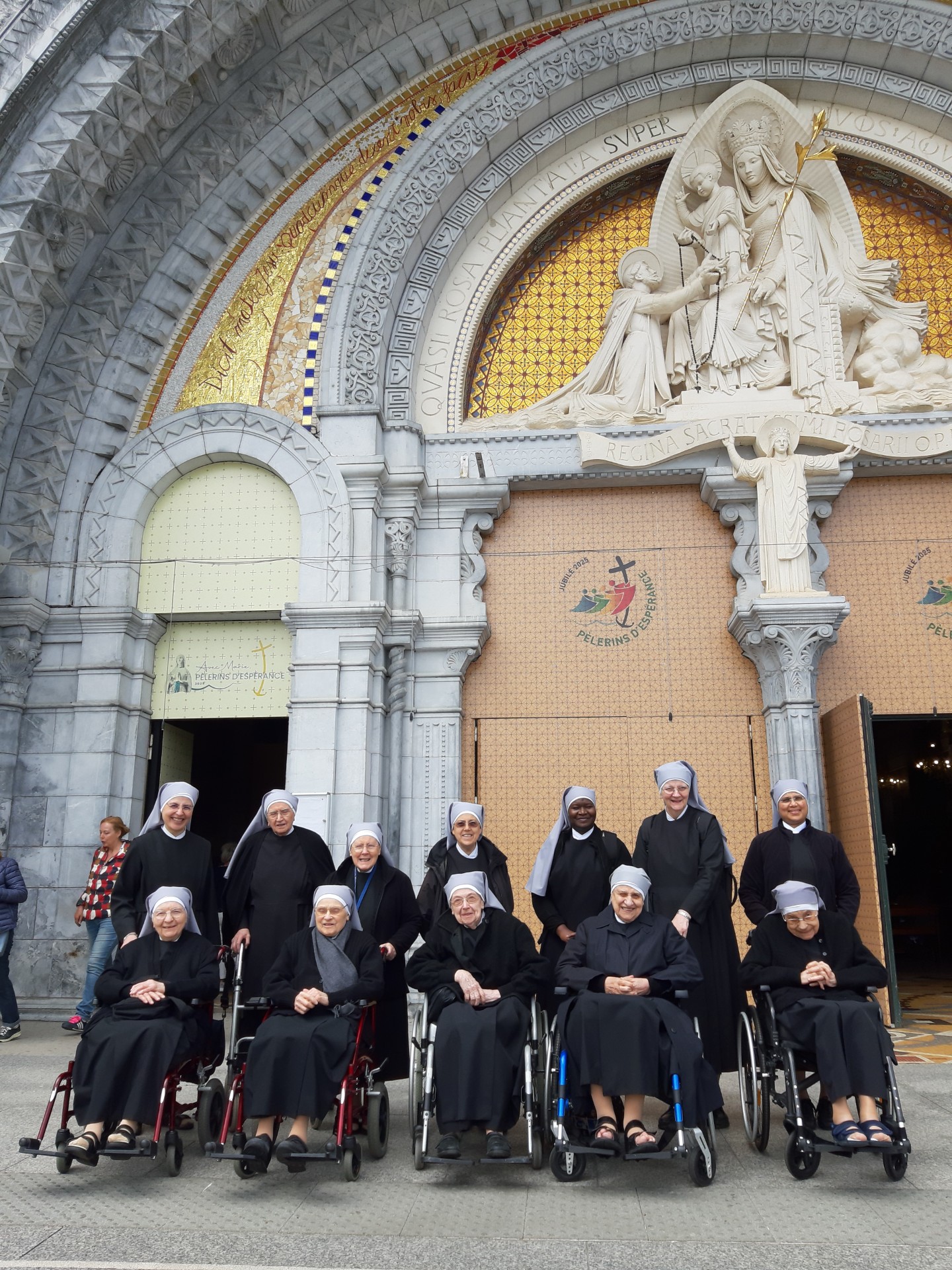 Petites Sœurs des Pauvres en fauteuil roulant et debout devant la basilique de Lourdes